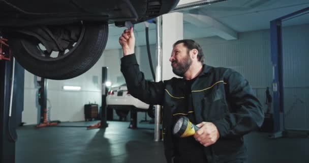 Homme mécanicien mature dans un centre de service automobile moderne tenant une lampe de poche, il fait un check-out de la voiture — Video