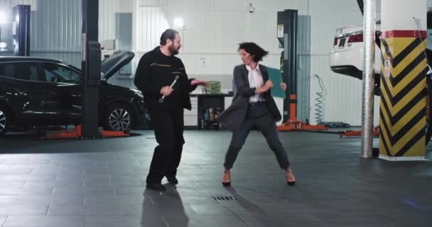 Señora guapa en un traje y hombre mecánico en el garaje de automóviles bailando excitado y divertido delante de la cámara — Vídeos de Stock