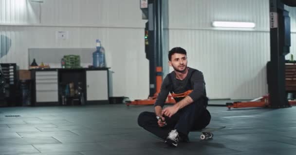 Snygg mekaniker i mitten av garaget sitter ner på skateboarden och leker med en skruvmejsel — Stockvideo