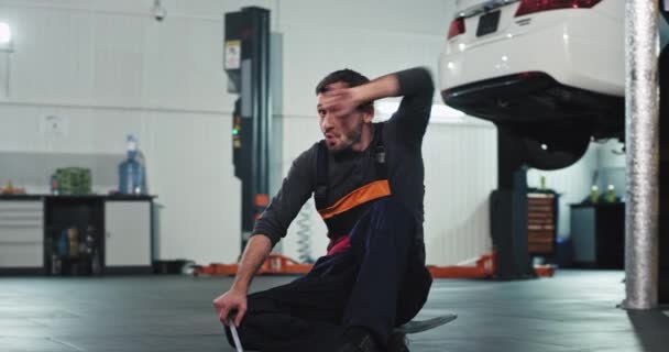 Porträtt av mekaniker man i uniform upphetsad framför kameran sitter ner på skateboard han känner sig trött efter jobbet — Stockvideo