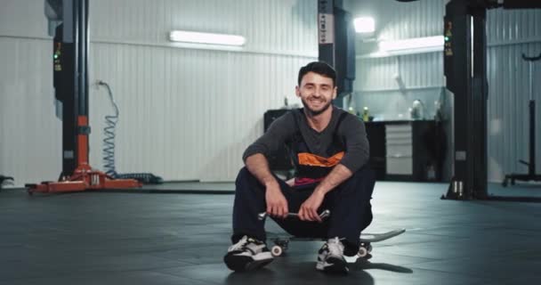 Portrait of a attractive mechanic guy in a uniform in front of the camera smiling large and showing big like he sitting down on the skateboard