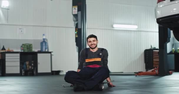 Chico mecánico bien parecido delante de la cámara sonriendo grande y sintiéndose emocionado en un el centro de servicio automático que tiene un sentarse en su monopatín — Vídeos de Stock