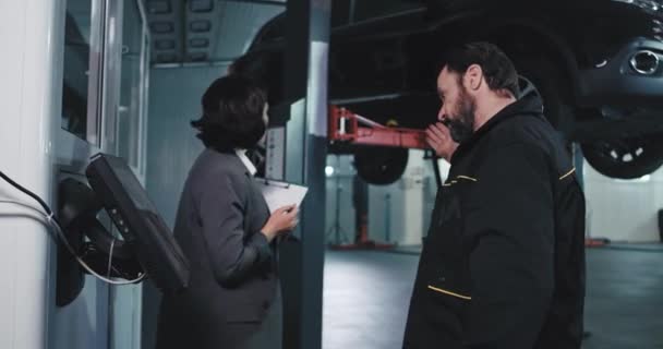 Mature mechanic man in the uniform have a conversation with the main manager from auto service center she check the plan of work and then they come under the damaged car to analyzing the problem of — Stock Video