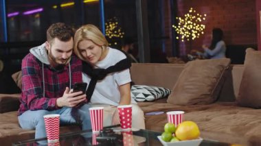 Sarışın, kısa saçlı ve karizmatik bir adam sırıtıyor. Kanepede oturup birlikte vakit geçiriyorlar. Telefondan bakıp sohbet ediyorlar.