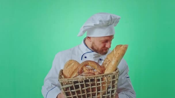 Piekarz w mundurze trzymający przed kamerą kosz świeżego chleba, wąchający chleb, kręcący film w zielonym tle studia. — Wideo stockowe