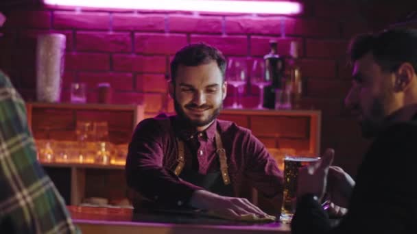 Garçom animado limpando a mesa de bar, enquanto dois caras cliente acelerando um grande momento em uma mesa de bar beber cerveja e desfrutar da atmosfera — Vídeo de Stock