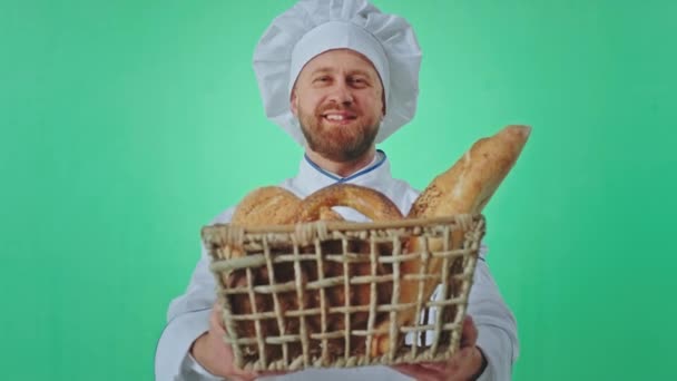 Sorridente grande panettiere in uno studio verde che tiene un grande cesto pieno di pane fresco e guarda dritto verso la fotocamera. Girato su ARRI Alexa Mini — Video Stock