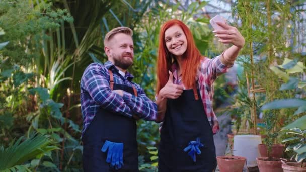 Atractiva jardinera hembra y hombre se toman selfies con un smartphone en un gran invernadero disfrutan del tiempo en su lugar de trabajo — Vídeos de Stock