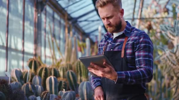 Framför kameran trädgårdsmästare man med hjälp av en tablett för att kontrollera tillståndet för växter i ett stort industriellt växthus tar han en telefon för att svara på samtalet — Stockvideo