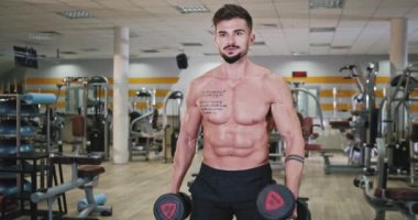 Spacious fitness studio athletic guy attractive looking in front of the camera professional doing the exercises for his biceps and triceps. 4k