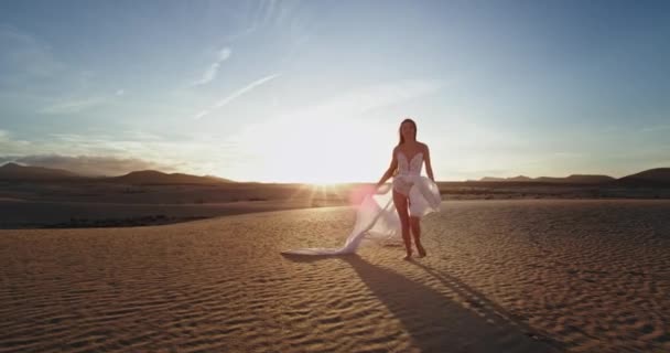 Красивая женщина с длинными волосами в белом платье медленно бежит по удивительному полю, полному песка перед камерой. выстрел по красному эпосу — стоковое видео