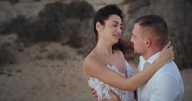 Beyaz elbiseli, mutlu ve gülümseyen bir çift harika bir manzaranın ortasında birbirlerine sarılırken çok güzel vakit geçiriyorlar.