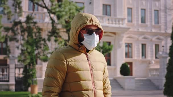 Человек в защитной маске на лице в желтой куртке посередине улицы в солнечных очках, смотрящих прямо в камеру, новый Ковид-19 — стоковое видео