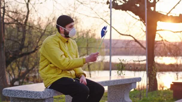 Parktaki genç adam gün batımında sandalyede oturuyor koruyucu bir maske takıyor ve Covid-19 Coronavirus salgınıyla oynuyor. — Stok video