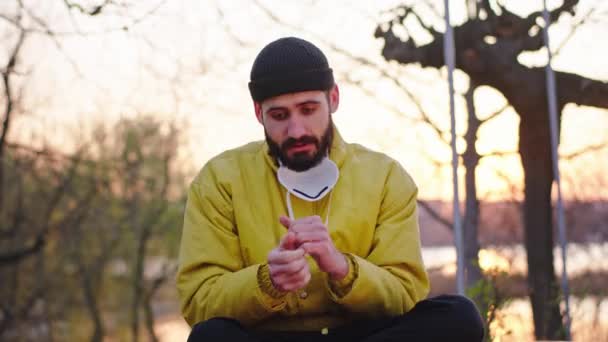 Un uomo freddo in mezzo al parco seduto sul chai al tramonto prende la maschera protettiva e si mette il viso in quarantena tempo Covid-19 N1H1 — Video Stock