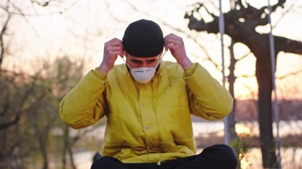 彼が一人で座って椅子に座っている間、彼の保護マスクを身に着けて日没で公園内の男は隔離されていますCovid-19.4k — ストック動画