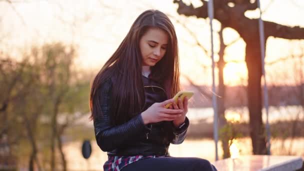 Krásná žena, která při západu slunce sedí v parku před kamerou a používá telefon, se soustředí — Stock video