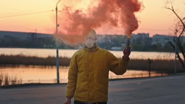 Atraktivní muž před kamerou uprostřed parku, držící kouřovou bombu, má na sobě ochrannou masku, dívající se přímo do kamery při západu slunce pandemie po celém světě Coronavirus 2019 — Stock video