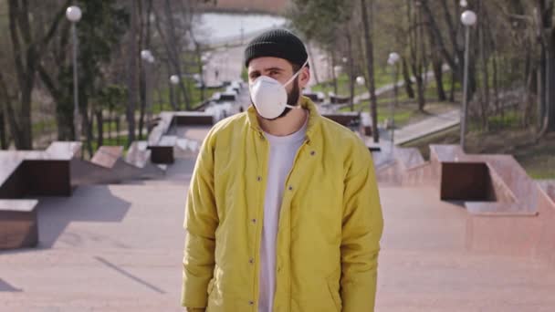 Um cara com um casaco amarelo em pé na frente da câmera no meio das escadas do parque ele usando uma máscara protetora, enquanto é o vírus fora ele olhando chateado em torno do fundo parque vazio — Vídeo de Stock