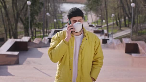 Parkın ortasında merdivenlerde koruyucu maskeli genç bir adam kameranın önünde telefonuyla konuşuyor boş bir park yeni Covid-19. — Stok video