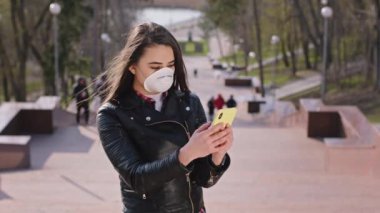 Korumacı maskeli, parkın yanındaki merdivenlerde kameranın önünde dikilirken telefonu kullanan karizmatik genç kadın, Covid-19 konsepti.