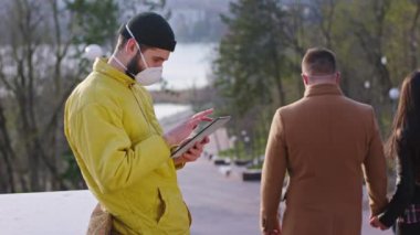 Yeni Covid-19 konsepti parkın ortasında sarı ceketli bir adam tablet kullanıyor koruyucu maskesi var arka planda merdivenlerden inen başka bir çift var.