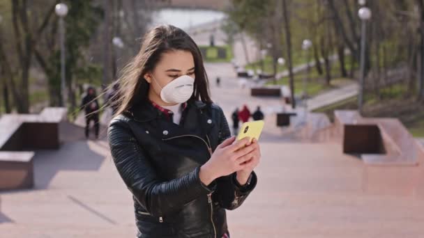 Korumacı maskeli, parkın yanındaki merdivenlerde kameranın önünde dikilirken telefonu kullanan karizmatik genç kadın, Covid-19 konsepti. — Stok video