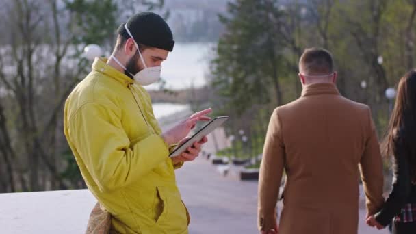Новая концепция Covid-19 в центре парка парень в желтой куртке с помощью планшета записывает что-то у него есть защитная маска другая пара на заднем плане прогуливаясь по лестнице у них есть — стоковое видео
