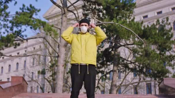 Um estudante com a jaqueta amarela que ele está no meio da rua vazia e tomar ele máscara protetora colocar em seu rosto e olhando ao redor novo Covid-19 — Vídeo de Stock