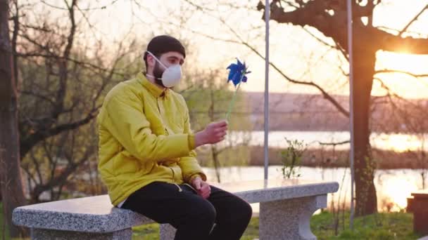 Konzept des gesunden und sicheren Lebens N1H1 ein Kerl sitzt auf dem Parkstuhl mit einer Schutzmaske auf dem Gesicht er spielt mit einem Windspinner er hat ein verärgertes Gesicht — Stockvideo