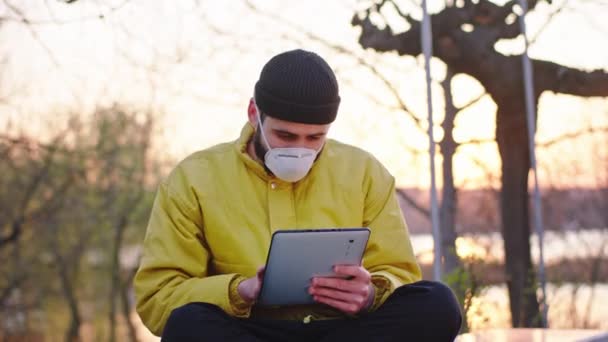 Συγκεντρωμένος άντρας με tablet ενώ κάθεται στην καρέκλα του πάρκου δακτυλογραφεί κάτι ενώ έχει μια προστατευτική μάσκα στο πρόσωπό του Coronavirus mers N1H1 — Αρχείο Βίντεο