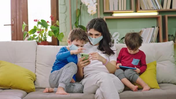 A kanapén anya a gyerekek viselik a védő maszk maradnak otthon, és használja az okostelefon játszani a játékot a kisfiú nem viseli a védő maszk karantén ideje új Coronavirus 2019 — Stock videók