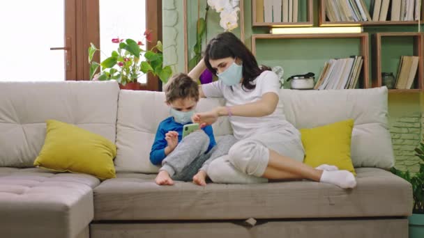I vardagsrummet karismatisk mor med sitt barn på soffan de bär skyddsmask leka tillsammans attraktiva spel på smartphone medan du bor hemma på karantän — Stockvideo