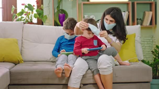 Nouvelle quarantaine Covid-19 à la maison jeune maman avec ses enfants prendre soin et pour la sécurité, elle porte le masque de protection pour les enfants assis sur le canapé — Video