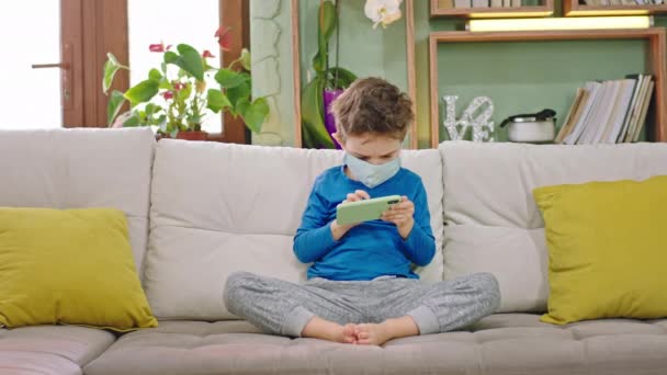 Дома в карантине 5-летний мальчик, играя сконцентрировал игру на своем смартфоне, сидя на диване концепции нового Covid-19 — стоковое видео