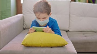 Akıllı telefonlu küçük bir çocuğun portresi kanepede koruyucu bir maskeyle oturup karantina Coronavirus 2019 salgınında konsantre bir oyun oynuyor.