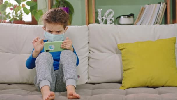 Дома в карантине сконцентрированный маленький мальчик в защитной маске играет в игры на смартфоне, сидя на диване Коронавирус 2019 — стоковое видео