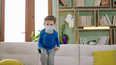 Küçük komik bir çocuk karantinada koruyucu maske takmış bir şekilde kanepede zıplarken oturma odasında hız yapıyor ve akıllı telefondan oynuyor.