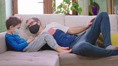 Evdeki kanepede küçük oğluyla birlikte koruyucu maske takarak vakit geçirirken oyun oynamak için akıllı telefon kullanıyorlar Coronavirus 2019