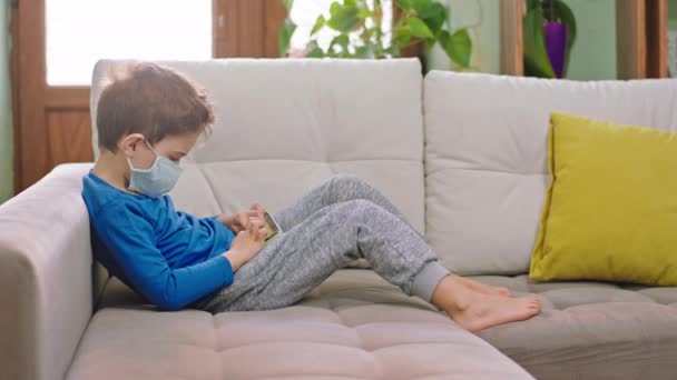 Med en skyddande mask liten pojke med en smartphone sitter på soffan och spela ett spel i karantän konceptet av en Covid-19 — Stockvideo