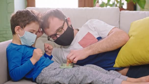 Un père avec son petit enfant reste à la maison en quarantaine ils jouent sur le smartphone tout en portant le concept de masque de protection de Coronavirus 2019 — Video