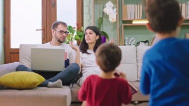 Oturma odasında iki çocuklu genç bir aile kanepede otururken iki küçük çocuk ellerinde kitaplarla ailelerine koşup masal okuyorlar.