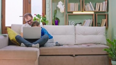 Genç baba ve küçük karizmatik oğlu kanepede mutlu vakit geçiriyorlar dizüstü bilgisayarda oynuyorlar ve evde birlikte vakit geçiriyorlar.