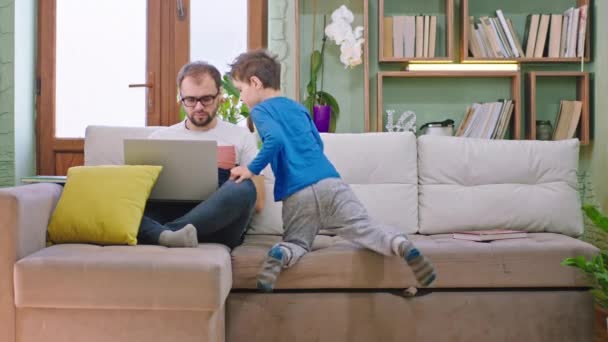 Vader werkt thuis op zijn laptop terwijl hij op de bank zit zijn zoontje komt naar hem toe zittend op de bank en begint te kletsen — Stockvideo