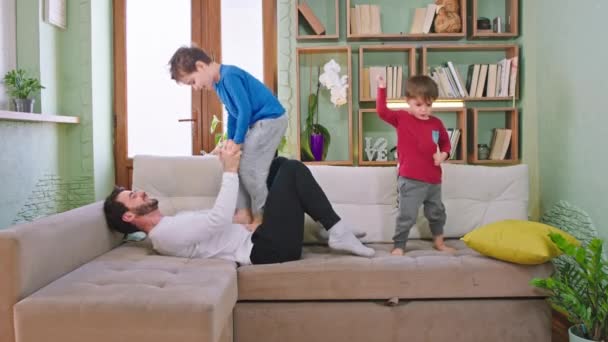 Hübsche und lustige kleine Kinder verbringen eine gute Zeit mit ihrem Vater, während sie sich auf das Sofa legen, auf dem sie kämpfen und die Zeit genießen — Stockvideo