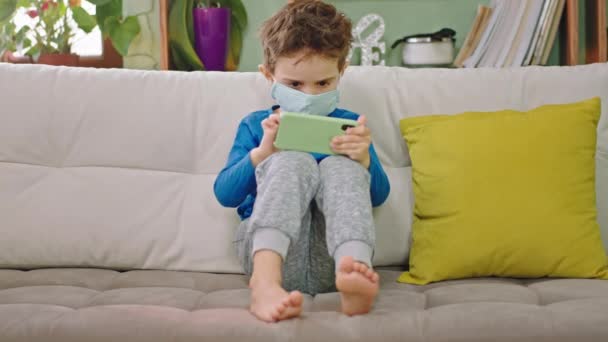 A kamera előtt karizmatikus fiú 5 éves koncentrált játszik a játékot az okostelefonján viseli a védő maszkot az arcán koncepció egy új Covid-19 — Stock videók