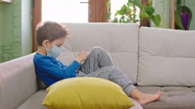 Coronavirus 2019 karantinaya alınıyor. Küçük çocuk, kanepedeki akıllı telefonu alıp oyun oynarken koruyucu maske takıyor.