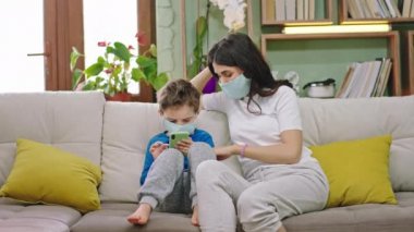 Kameranın önünde çekici bayan anne ve çocuğu evdeki kanepede oturuyor akıllı telefonu oyun oynamak için kullanıyor anne ona yeni Coronavirus 2019 karantinasında yardım ediyor.