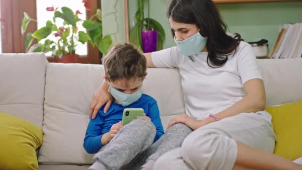 Kameranın önünde annesinin koruyucu maskesini takan tatlı bir çocuk kanepede otururken annesi çocukla vakit geçirip akıllı telefonu oyun karantinası için kullanıyor. — Stok video