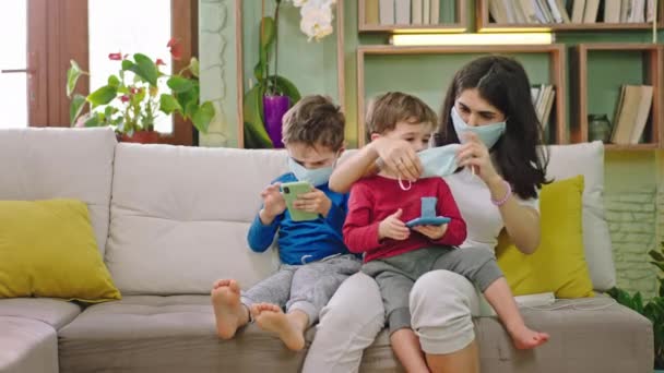 Thuis in quarantaine beschermt de jonge moeder haar kinderen met het beschermend masker op hun gezicht terwijl ze het spel spelen op het smartphone concept van een nieuwe Covid-19 — Stockvideo